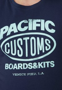 Granatowa bawełniana koszulka z białym, postarzałym napisem "PACIFIC CUSTOMS BOARDS&KITS VENICE PIER, L.A." w odważnej, zaokrąglonej czcionce.