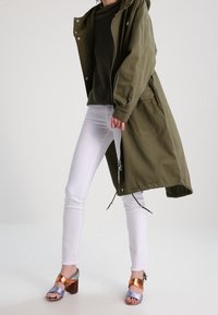 Cappotto lungo verde oliva con cappuccio, indossato sopra un maglione scuro. Jeans skinny bianchi e sandali con tacco a blocco bicolore con cinturini in rame e blu.