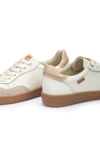 Zapatilla de cuero blanca con acentos de ante beige, puntera redonda, cordones color crema y suela de goma. Presenta detalles de costura y una etiqueta con el logotipo en el lateral.