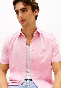 Ροζ πουκάμισο με κοντά μανίκια και γιακά με κουμπιά, φτιαγμένο από βαμβάκι, με μικρό κεντημένο λογότυπο στο στήθος, φορεμένο πάνω από λευκή φανέλα.