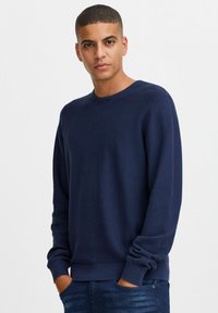 Marineblå, strukturert genser med rund hals og ribbede mansjetter, kombinert med tettsittende jeans. Tøyet har et subtilt mønster.