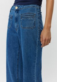 Korkeavyötäröiset tummansiniset farkut, joissa on leveät lahkeet, kahdella takataskulla ja hienovaraisella ompeluyksityiskohdalla. Sileä pinta ja klassinen muotoilu.