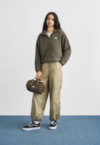 Πουλόβερ από φλις σε ελαιόχρωμο με φερμουάρ μέχρι τη μέση, συνδυασμένο με μπεζ cargo παντελόνι με πλαϊνές επιφάνειες, μαύρα και άσπρα αθλητικά παπούτσια και καφέ τσάντα duffel.