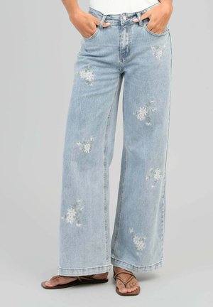 Jean bleu clair à jambes larges avec broderie florale blanche, porté avec des sandales marron à lanières et les mains dans les poches.