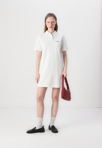 Biela polo šaty s krátkymi rukávmi, golierom a vyšívaným logom. Čierne mokasíny, svetlošedé ponožky a červená kabelka na rameno dotvárajú outfit.