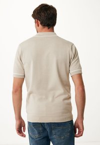 Béžové polo tričko s krátkým rukávem a texturovaným pleteným povrchem, s bílým lemem na límci a rukávech, v kombinaci s modrými džíny.