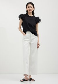 T-shirt nera con maniche in tulle arricciato, abbinata a pantaloni bianchi a gamba larga e sandali neri a scivolo. Design pulito e moderno con colori a contrasto.