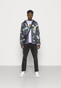 Giacca camouflage in verde scuro e grigio, sopra una maglietta viola chiaro con una grafica. Pantaloni neri, sneakers bianche e occhiali da sole arancioni completano il look.