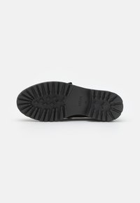 Suela de bota de goma negra con un patrón de relieve profundo, diseñada para ofrecer agarre. Presenta acentos geométricos elevados y la marca en el área del arco.