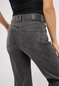 Grå denimjeans med en tætsiddende talje, to baglommer og et mærketag. Stoffet har en subtil tekstur med en let falmet overflade.
