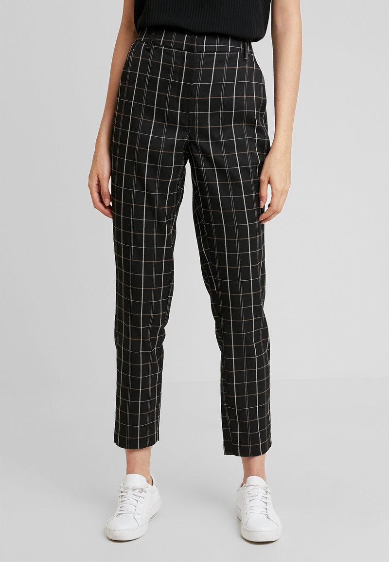 Personne portant un pantalon noir ajusté à carreaux fenêtres blancs, longueur cheville, et des baskets blanches, debout devant un fond uni.