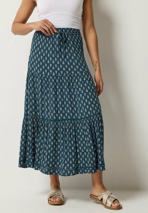 Kobieta w turkusowej, wzorzystej maxi spódnicy z wielowarstwowym fasonem, białym bezrękawniku i beżowych, plecionych sandałach wsuwanych na tle jednolitego tła.