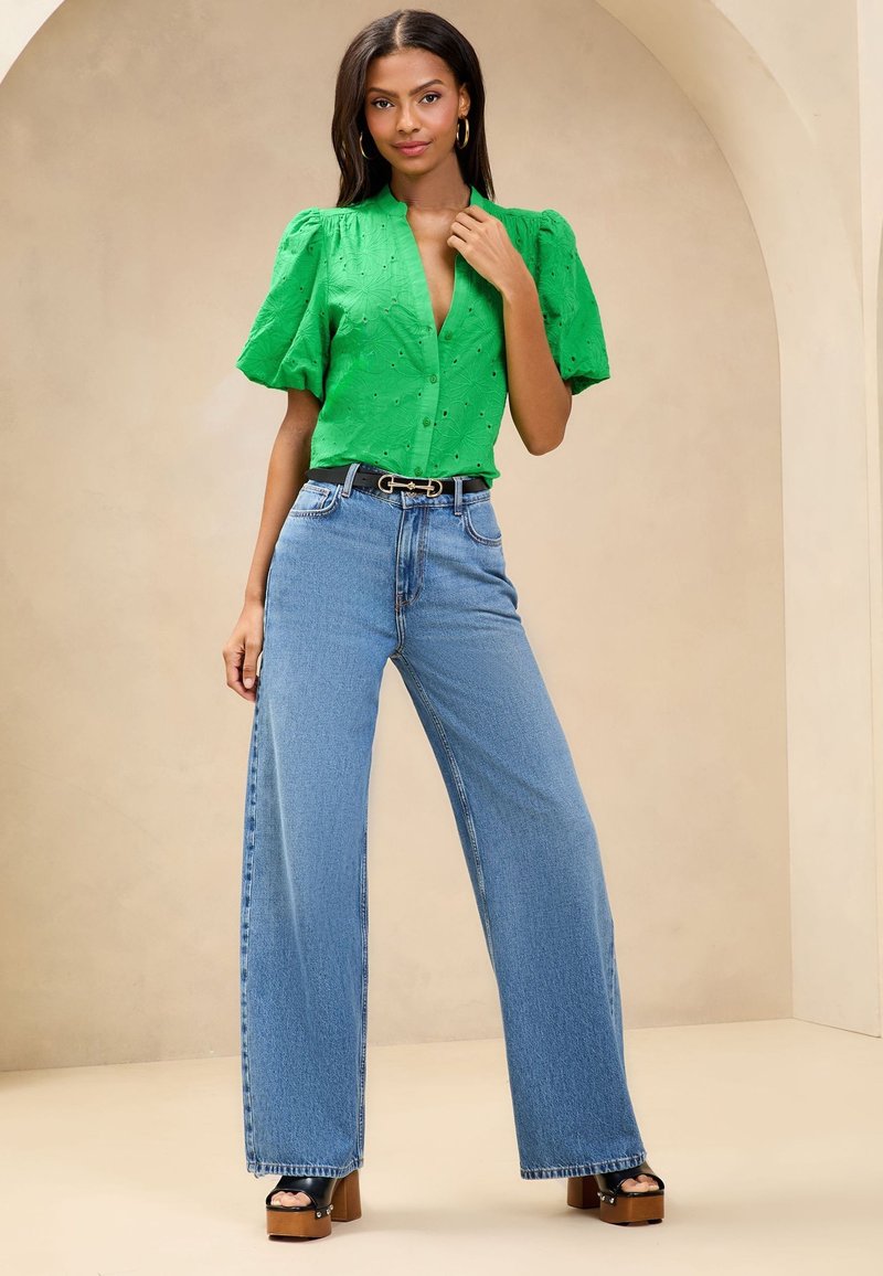 Donna con blusa verde brillante a maniche a sbuffo, jeans blu a vita alta e gamba larga, cintura nera e sandali neri con plateau in piedi sotto un arco neutro.