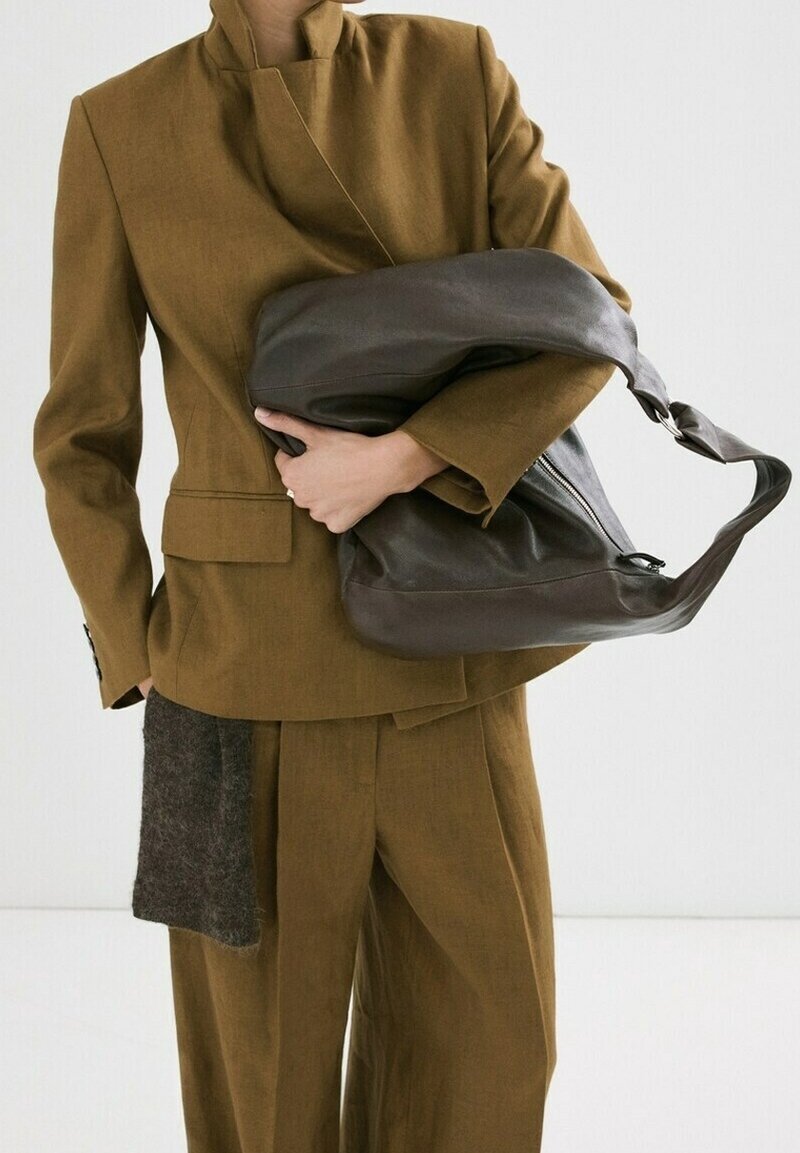 Personne portant un costume marron tenant un grand sac à bandoulière en cuir marron foncé sur fond blanc.