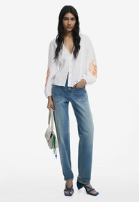 Weißes Langarmhemd mit orangefarbenen floralem Stickerei, kombiniert mit hellblauen hochgeschnittenen Jeans. Die Schuhe sind blaue Riemchenabsätze.