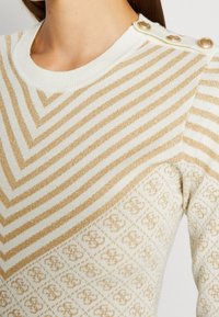 Gestrickter Pullover in Creme mit goldfarbenen diagonal verlaufenden Streifen und einem strukturierten Muster. Verfügt über goldene Knöpfe an der Schulter. Weicher, elastischer Stoff.