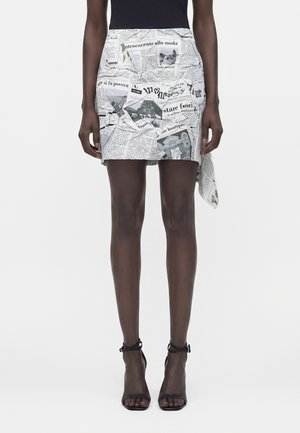 Personne portant une mini-jupe blanche avec un imprimé journal noir et blanc, associée à des talons hauts noirs à lanières et un haut noir.