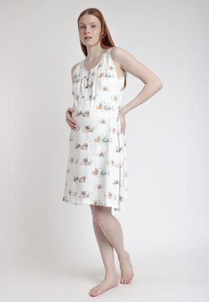 Mujer embarazada de pie, descalza, vestida con un camisón blanco sin mangas con estampados coloridos de animales de dibujos animados.