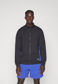 Svart idrettsjakke med blå sidepaneler, glidelås foran og høy krage. Har en hvit Puma-logo på venstre side. Kombinert med blå shorts.