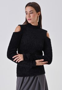 Donna che indossa un maglione nero peloso con spacchi sulle spalle e grandi orecchini a cerchio dorati, con le braccia incrociate, che guarda di lato su uno sfondo semplice.