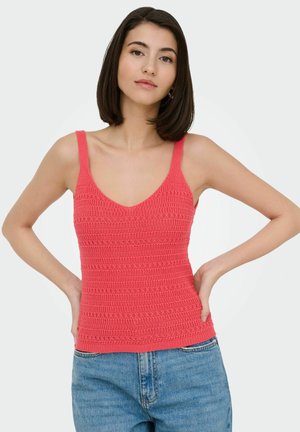 Woman with straight dark hair wearing a sleeveless coral knit top and blue jeans, posing with hands on hips against a plain background.