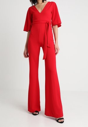 Femme portant une combinaison rouge vif avec un pantalon à jambes larges, un profond décolleté en V, des manches jusqu'aux coudes et une ceinture nouée à la taille.