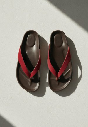 Brune slip-on sandaler med tykke røde stofstropper liggende side om side på en hvid overflade med sollys, der kaster skygger.