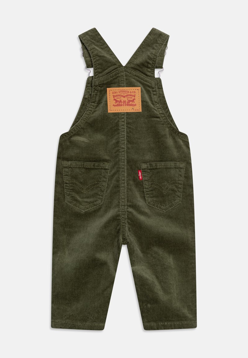 Pantalones de corduroy en verde olivo con un parche cuadrado en la parte trasera, tirantes ajustables y bolsillos frontales. Detalles de costura notables en los bolsillos.