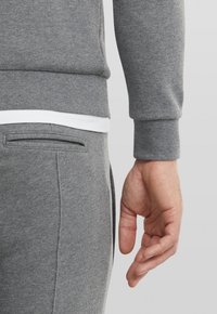Felpa grigia con orlo a coste bianco e pantaloni grigi abbinati. Caratteristiche includono una tasca laterale e tessuto testurizzato. Mano visibile di lato.