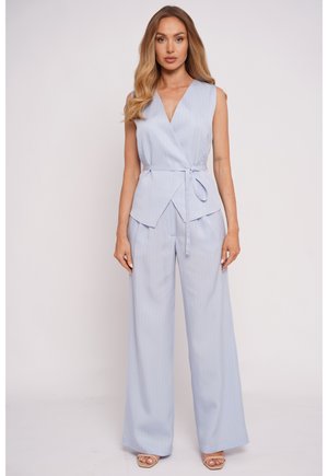 Kobieta ubrana w jasnoniebieską, bezrękawną bluzkę z kopertowym dekoltem oraz dopasowane spodnie z szerokimi nogawkami i beżowe sandały, stojąca na jednolitym tle.