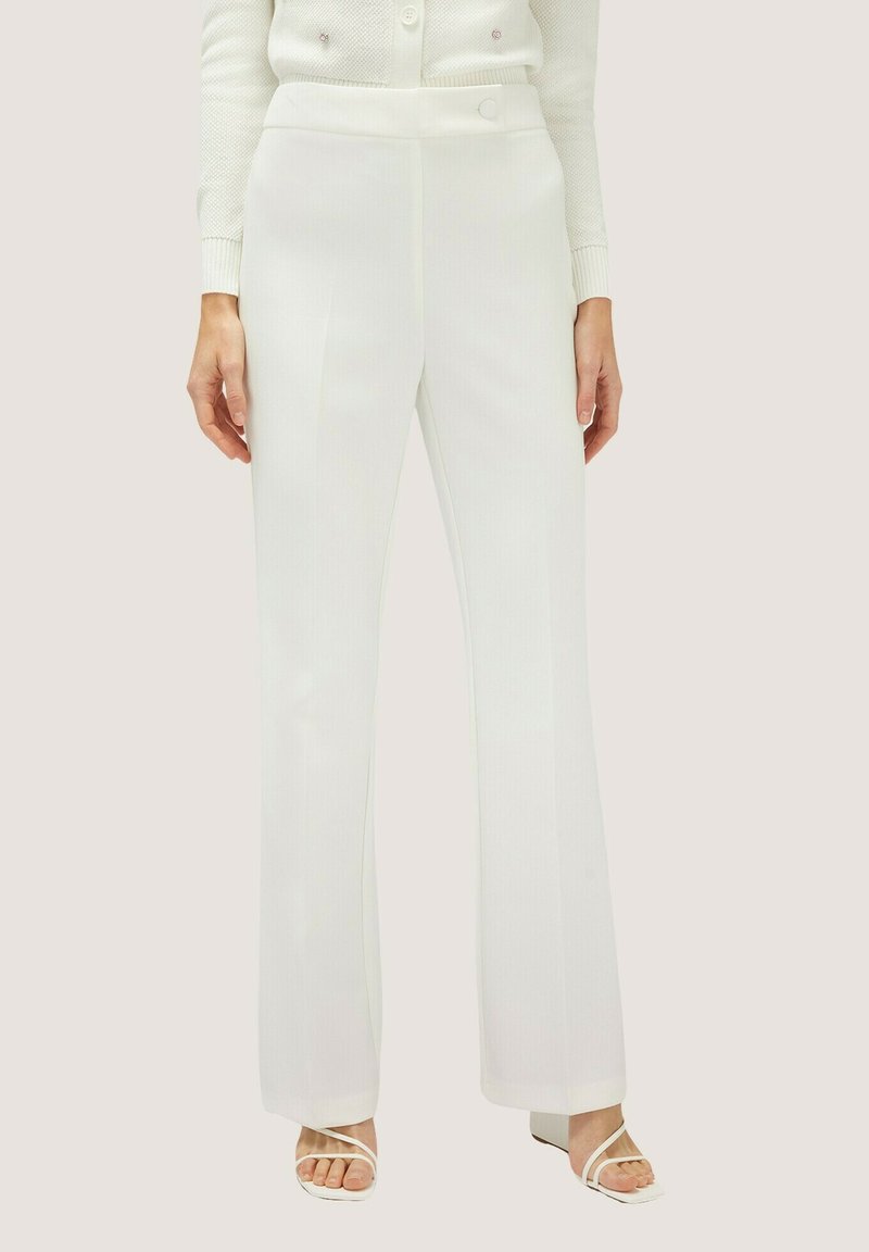 Pantaloni bianchi su misura con gamba dritta, texture liscia, vita alta e discreta fantasia a righe sottili, abbinati a semplici sandali bianchi.