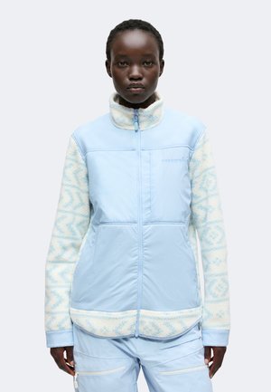 Persona que lleva una chaqueta azul claro con cremallera y mangas de forro polar blancas y azules con estampado, y pantalones cargo a juego en azul claro.