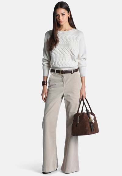 Femme portant un pull en maille torsadée blanc, un pantalon beige à jambes larges, une ceinture marron et tenant un sac à main marron avec des détails de franges.
