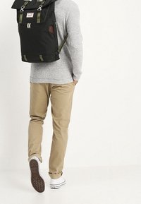 Czarny plecak z tkaniny, z zielonymi paskami, metalowymi elementami i klapą na górze, noszony przez osobę w jasnych spodniach i butach.