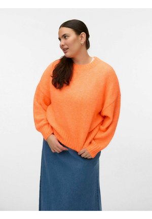 Vrouw met lang donker haar in een oversized feloranje trui en een blauwe denim rok, staand en zijwaarts kijkend tegen een effen witte achtergrond.