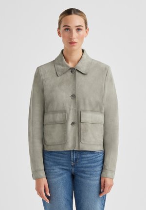 Frau trägt eine hellgraue Wildlederjacke mit Knöpfen, zwei Vorderklapptaschen und blaue Jeans, steht vor einem schlichten weißen Hintergrund.