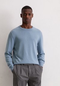 Jasnoniebieski dziany sweter z okrągłym dekoltem, noszony na szarych plisowanych spodniach. Gładka tekstura, dopasowane rękawy oraz subtelny detal z logo.