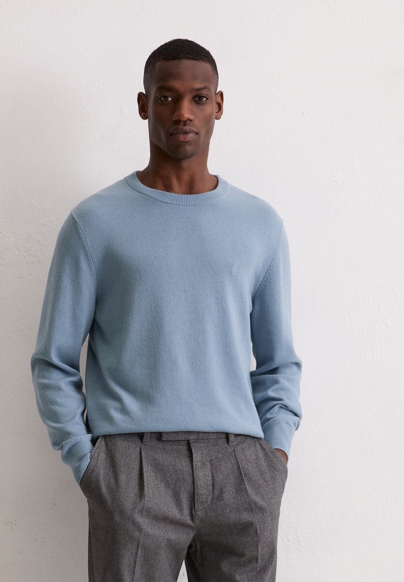 Jasnoniebieski dziany sweter z okrągłym dekoltem, noszony na szarych plisowanych spodniach. Gładka tekstura, dopasowane rękawy oraz subtelny detal z logo.