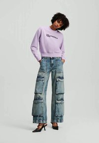 Mulher vestindo um sweatshirt lilás da Karl Lagerfeld, calças cargo de denim largas com vários bolsos e sapatos de salto pretos, em pé com as mãos nos bolsos.