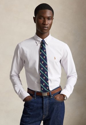 Mand iført en hvid skjorte med knapper, blåstribet slips, brunt bælte, blå jeans og armbåndsur, stående med hænderne i lommerne mod en neutral baggrund.