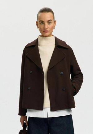 Vrouw met haar naar achteren gebonden, draagt een donkerbruine dubbelgeknoopte jas over een beige coltrui en houdt een zwarte handtas vast.