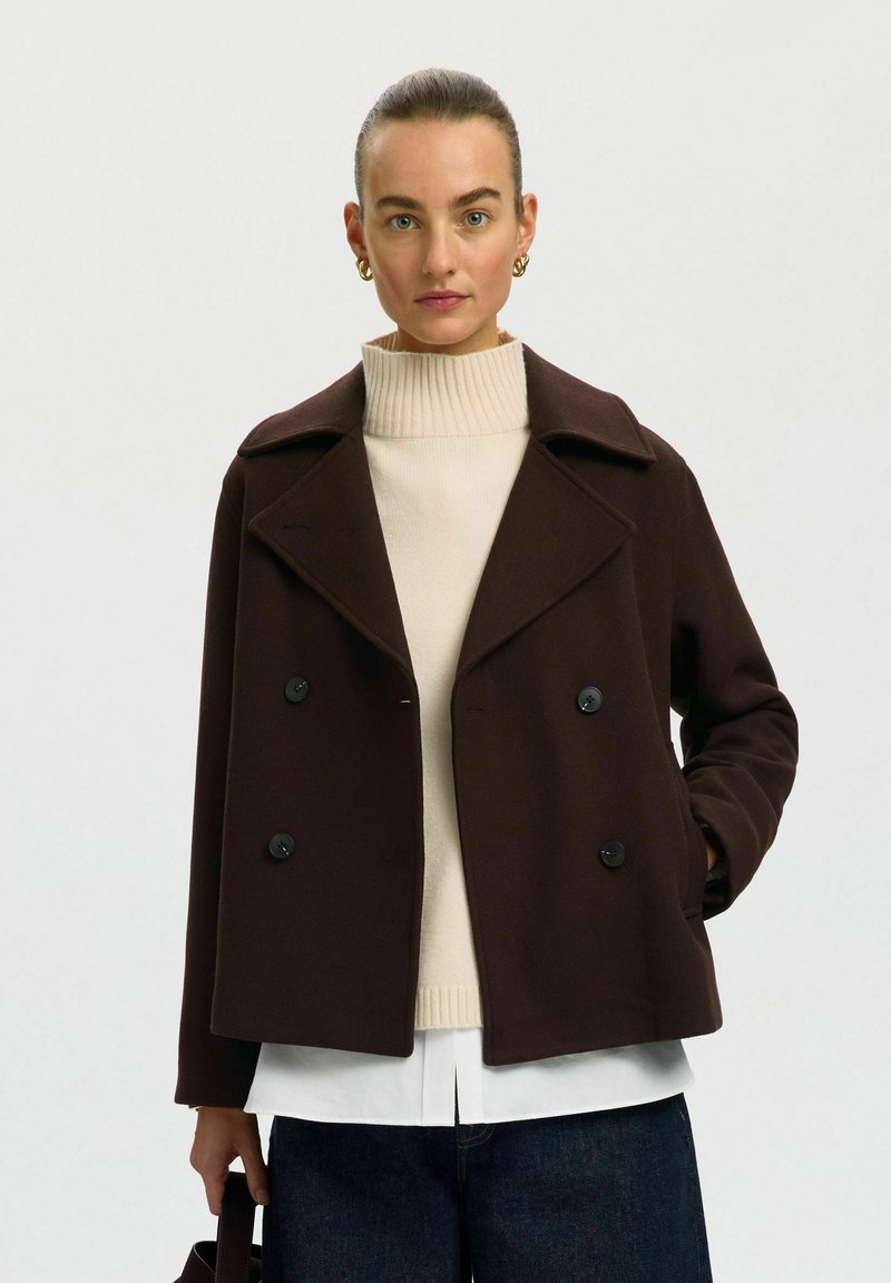 Femme aux cheveux attachés portant un manteau croisé marron foncé sur un pull à col roulé beige et tenant un sac à main noir.