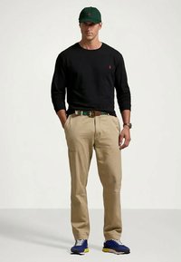 Homem a usar uma camisa de manga longa preta, calças caqui com cinto, boné verde, ténis azuis e relógio, de pé com as mãos nos bolsos.