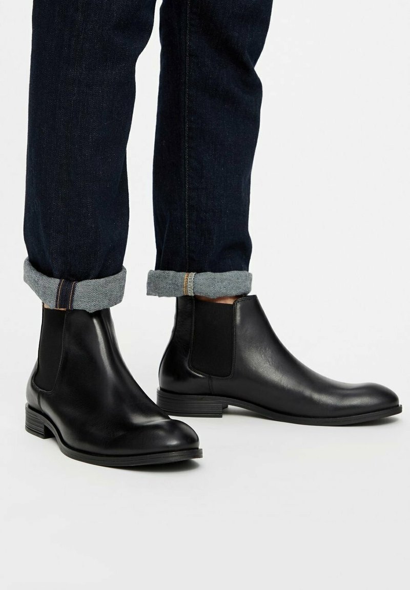 Bottes Chelsea en cuir noir portées avec des jeans foncés retroussés sur un fond blanc.