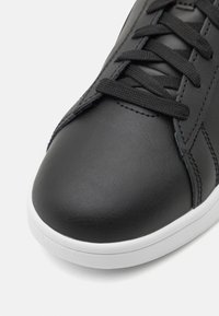 Zbliżenie czarnego skórzanego sneakersa z czarnymi sznurówkami i białą gumową podeszwą na białym tle.