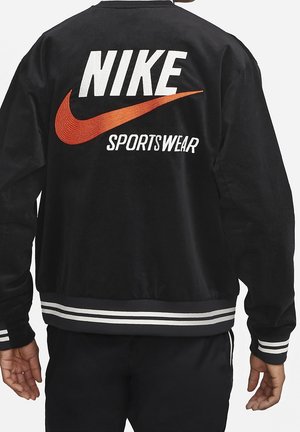 Personne portant une veste noire avec le logo Nike blanc et rouge et le texte "Sportswear" dans le dos, avec des poignets et un ourlet rayés blancs.