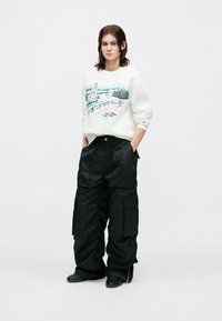 Witte sweatshirt met een landschap print in groen en roze, gecombineerd met oversized zwarte cargobroeken met grote zijzakken. Zwarte schoenen.