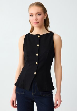 Donna che indossa un top nero senza maniche con bottoni dorati sul davanti, abbinato a jeans blu scuro, in piedi davanti a uno sfondo semplice.