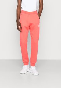 Korallfärgade sweatpants med elastisk midja, sidofickor, smalnande ben och vitt logodetalj. Matchas med vita sportskor.