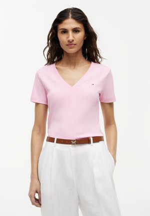 Ženska, ki nosi svetlo roza majico z V-izrezom, zataknjeno v bele hlače z visokim pasom in rjavim pasom, stoji pred belim ozadjem.